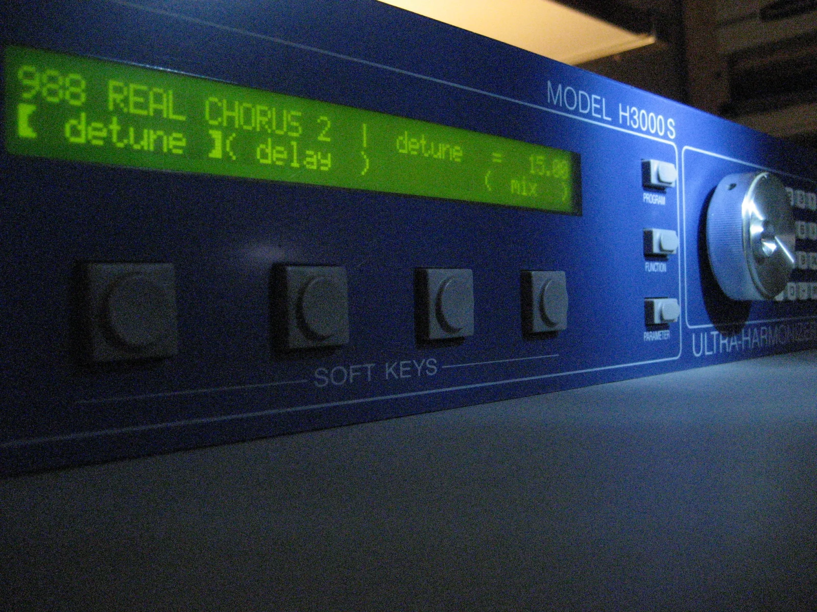 An angled view of the blue front panel. The green-backlit LCD shows “988 REAL CHORUS 2” and the detune and delay soft-key functions. Four soft keys sit below it, with three menu buttons and a knob on the right.